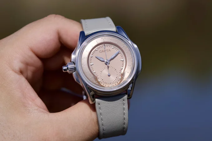 Hand holding a CAPTA watch with rose gold dial, sub-dial, silver case, and grey leather strap, set outdoors.