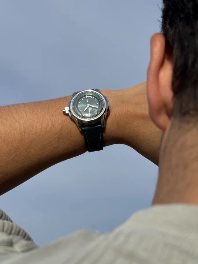 Man's arm displays a luxury watch: silver case, speckled dark grey dial, green leather strap, under a blue sky.