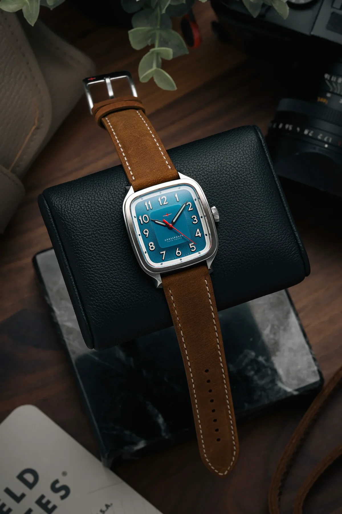 Cushion silver watch with teal dial & brown suede strap, on a black leather watch roll, camera & plant nearby.
