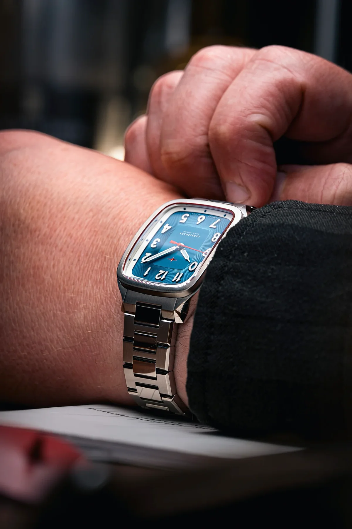 Close-up of a wrist wearing a silver tonneau watch with a blue dial, white numerals, red second hand, and steel bracelet.