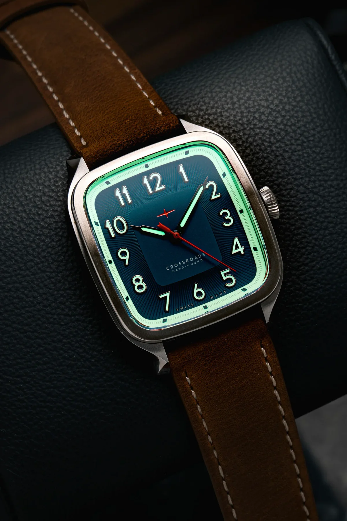 Cushion-cased steel watch with glowing green bezel, dark teal dial, luminous numerals, red seconds hand, brown suede strap on a black surface.