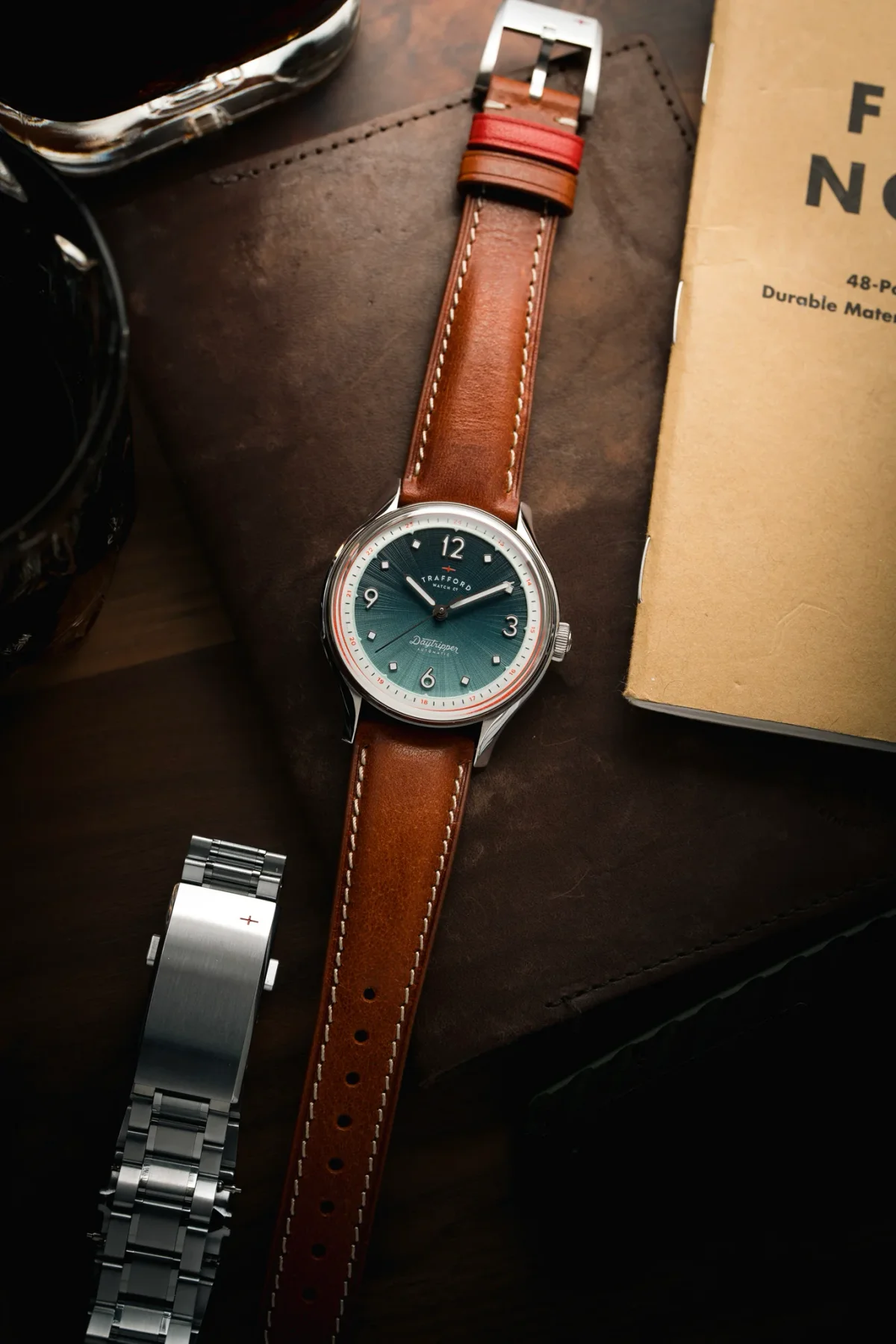 Trafford Daytripper watch with green gradient dial, brown leather strap with red accent, steel bracelet, on dark leather with notebook.