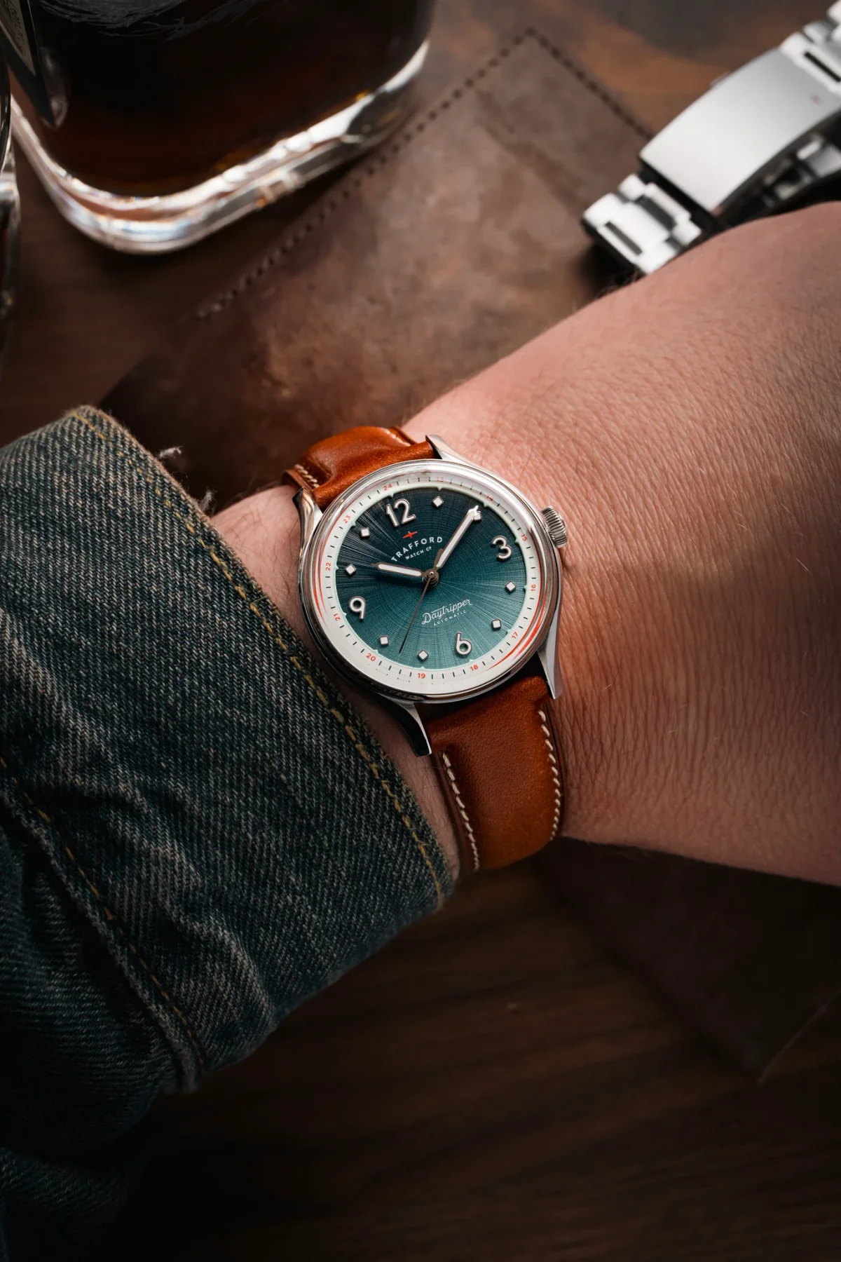 Wrist with Trafford Daytripper Automatic watch: teal sunburst dial, brown leather strap. Denim sleeve, blurred glass & steel bracelet.
