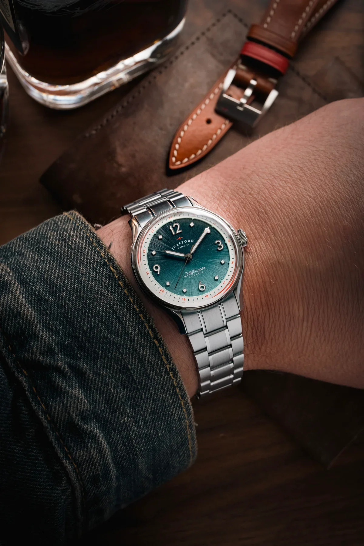 Wrist wearing a Trafford watch with green sunburst dial, steel bracelet, and denim cuff. Decanter & leather strap visible.