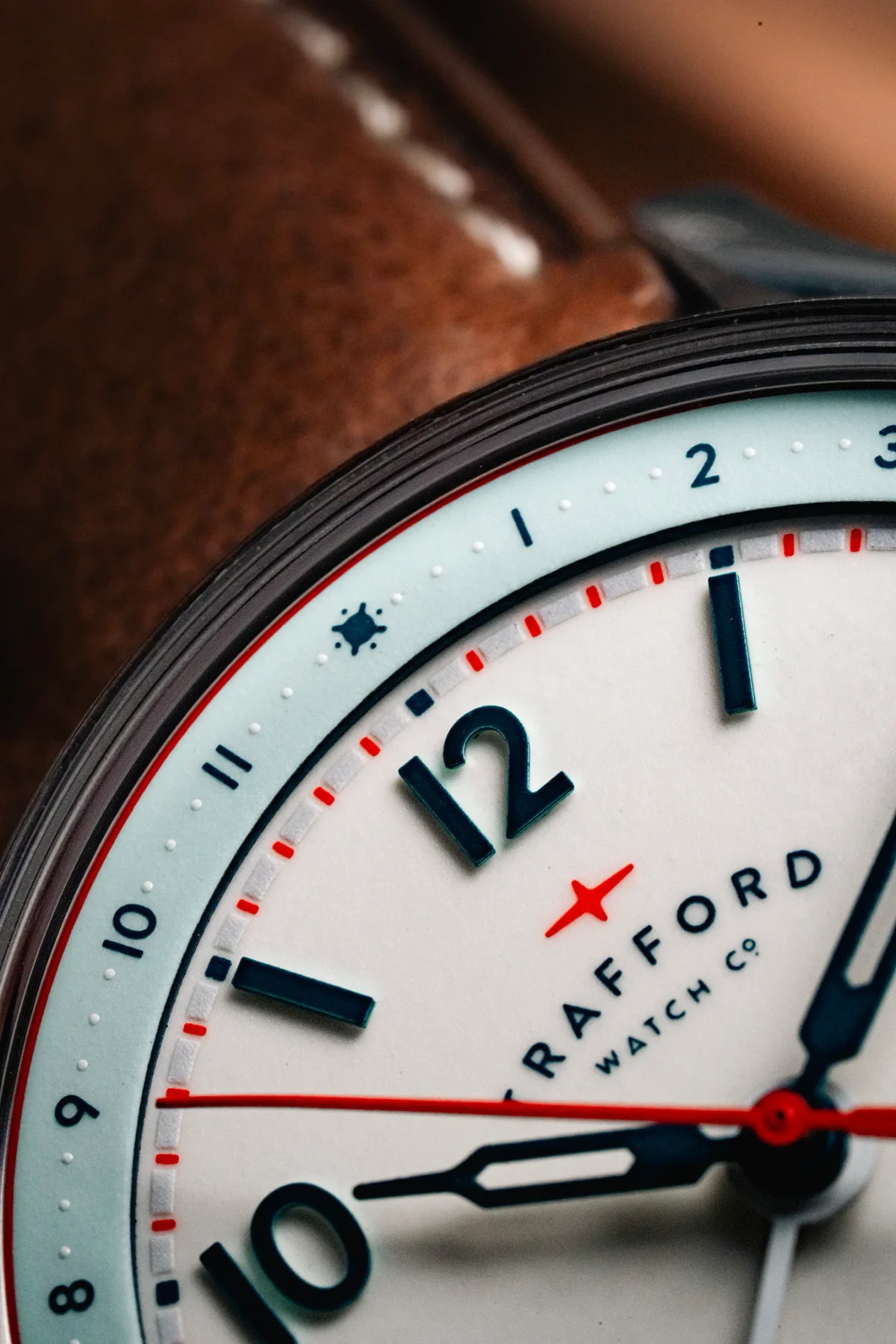 Close-up of a Crafford watch dial. White face, blue numbers & hands, red accents, light blue outer ring. Brown leather background.
