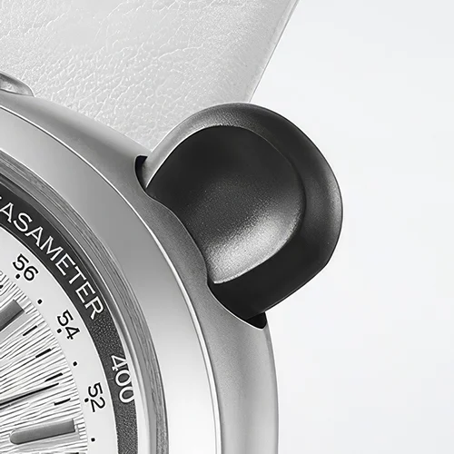 Close-up of a silver watch case featuring a prominent black rounded pusher. Part of a tachymeter dial and a white strap are visible.