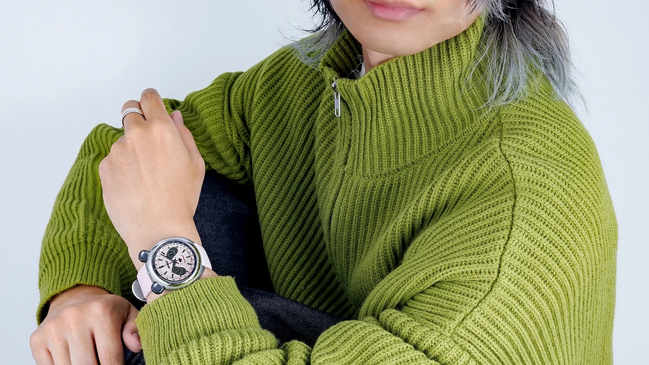 Person in green sweater wears a Seiko panda-faced chronograph watch with black 'ears', white dial, and pink strap.