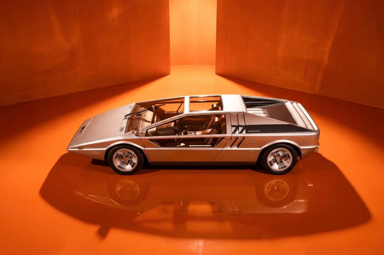 Silver 1972 Maserati Boomerang concept car with angular lines, in a vibrant orange room on a reflective floor.