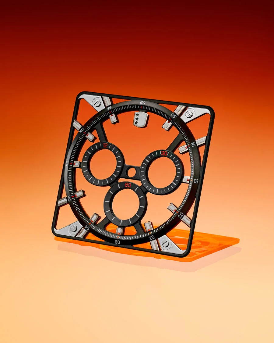 Skeletal black watch component with square silver frame, three chrono subdials, red accents on an orange gradient.