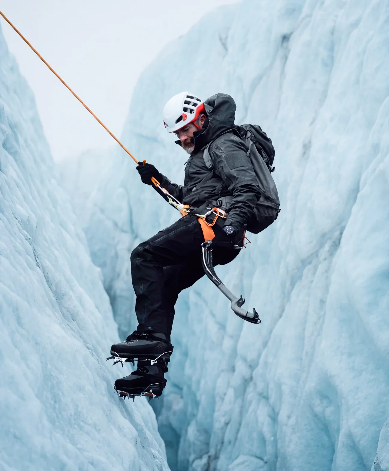 Man in full climbing gear, helmet, crampons & ice axe, rappelling down blue glacial crevasse, watch on wrist.