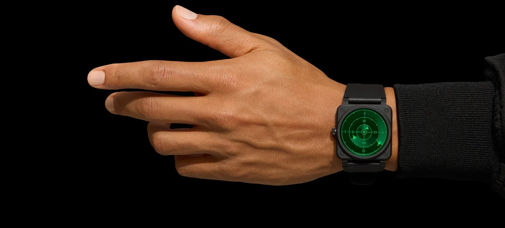 Brown hand pointing, wearing a square black watch with a green radar dial against a black background.
