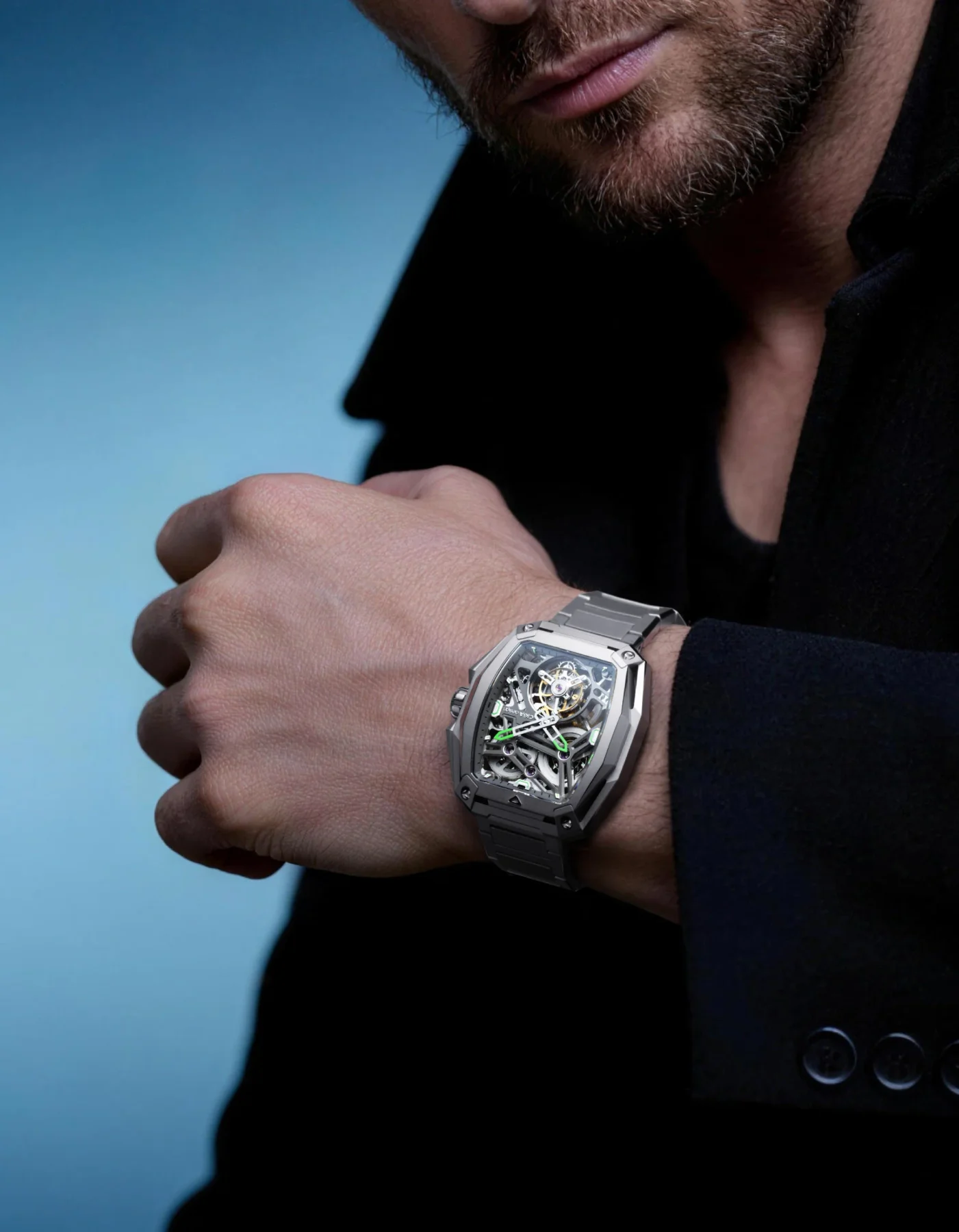 Man's bearded face & arm wearing a metallic skeleton watch with green accents & metal bracelet, against blue background.