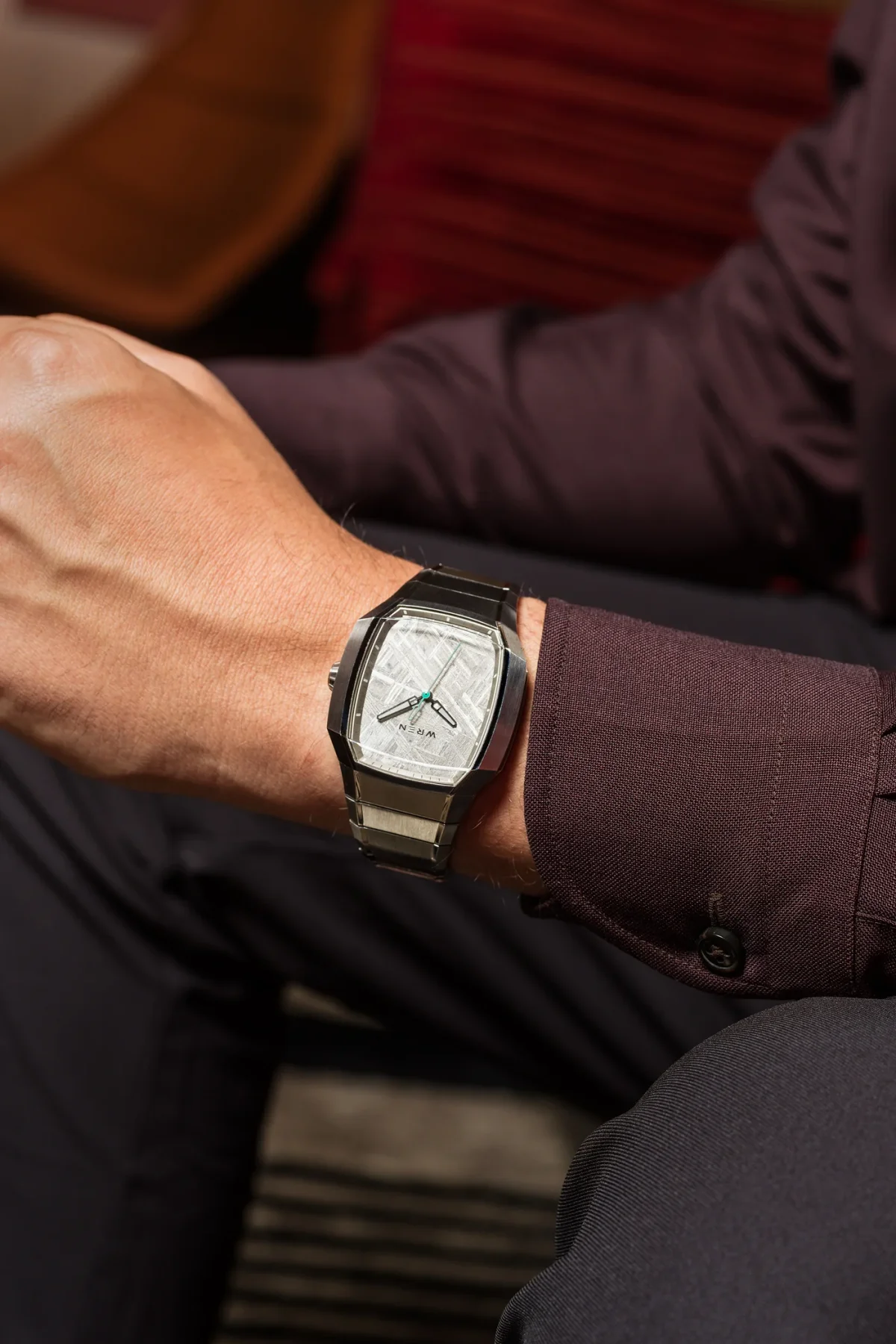 Man's arm wearing a modern steel Wren watch with a textured meteorite dial and teal seconds hand, plum shirt.