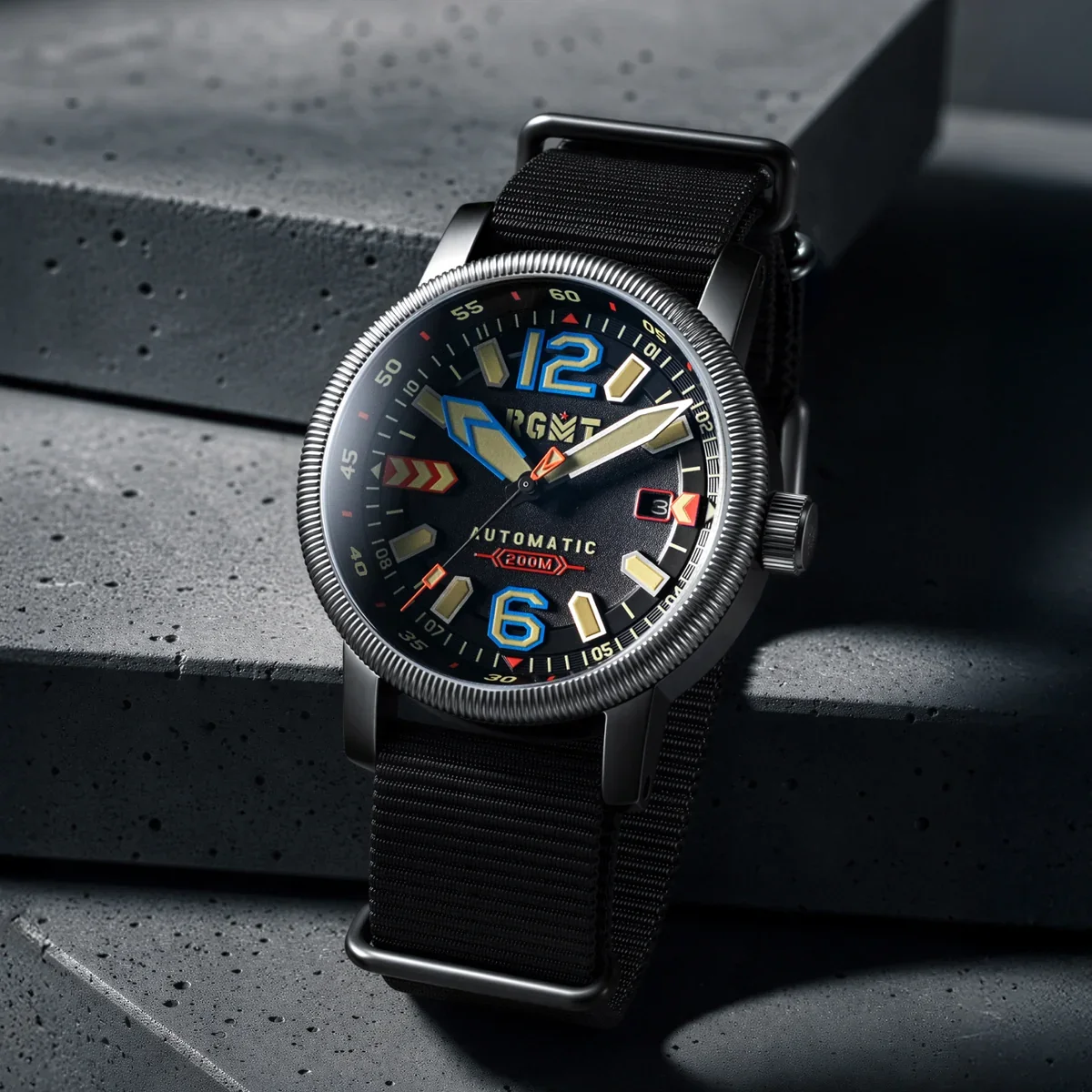 RGMT automatic watch with colorful dial on black NATO strap, fluted bezel, resting on a dark concrete surface.