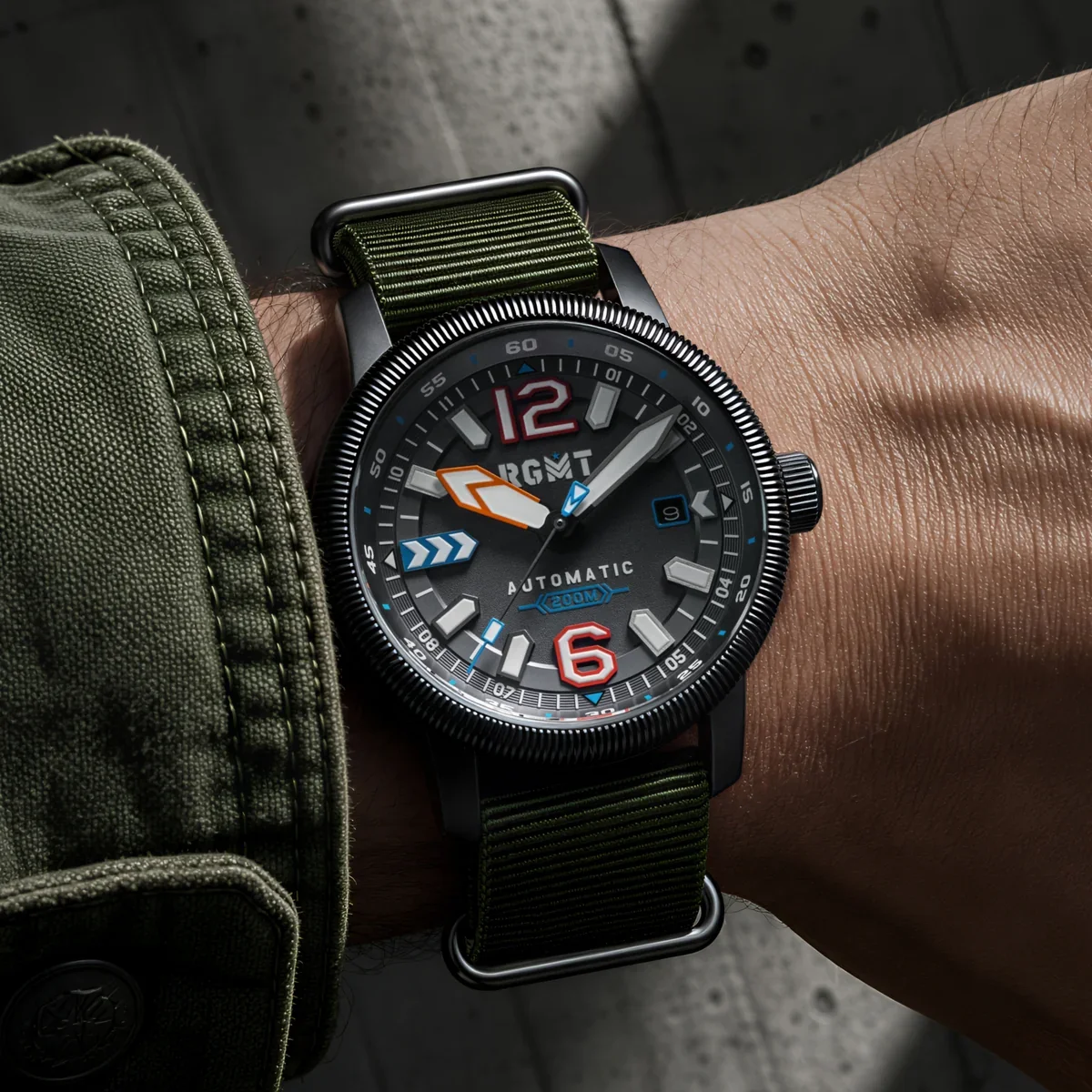 RGMT automatic watch on wrist: dark dial with red 12/6, colorful markers, orange arrow hand, and olive green NATO strap.