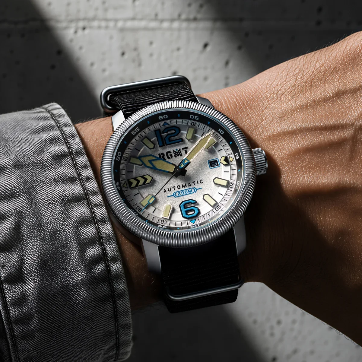 Wrist wearing RGMT automatic watch with silver dial, blue accents, ridged bezel, black NATO strap, against a concrete wall.