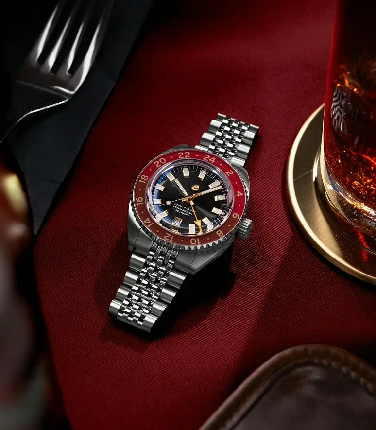 Steel GMT watch with red/black bezel, jubilee bracelet, on red cloth with glass of drink, fork.