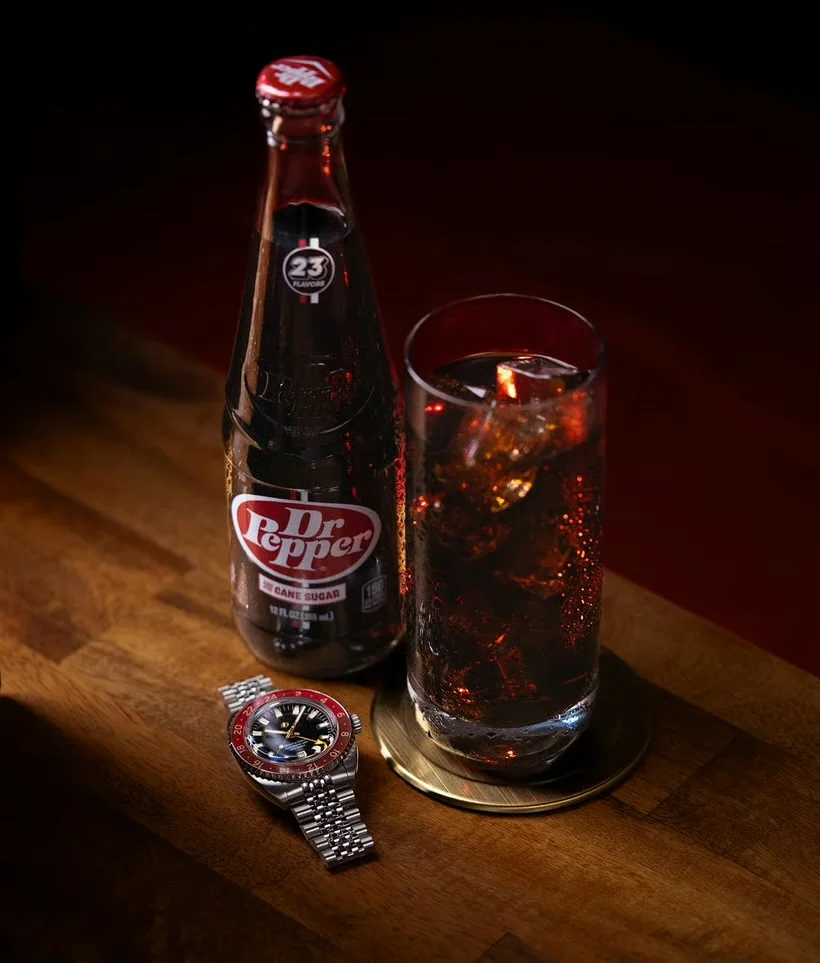 Luxury watch with black dial, red/blue bezel, jubilee bracelet next to Dr. Pepper bottle and glass on wood.