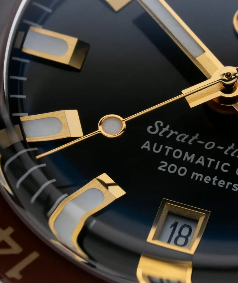 Extreme close-up of Strat-o-time watch dial: dark blue face, gold hands, luminous markers, date 18, 200m rating.