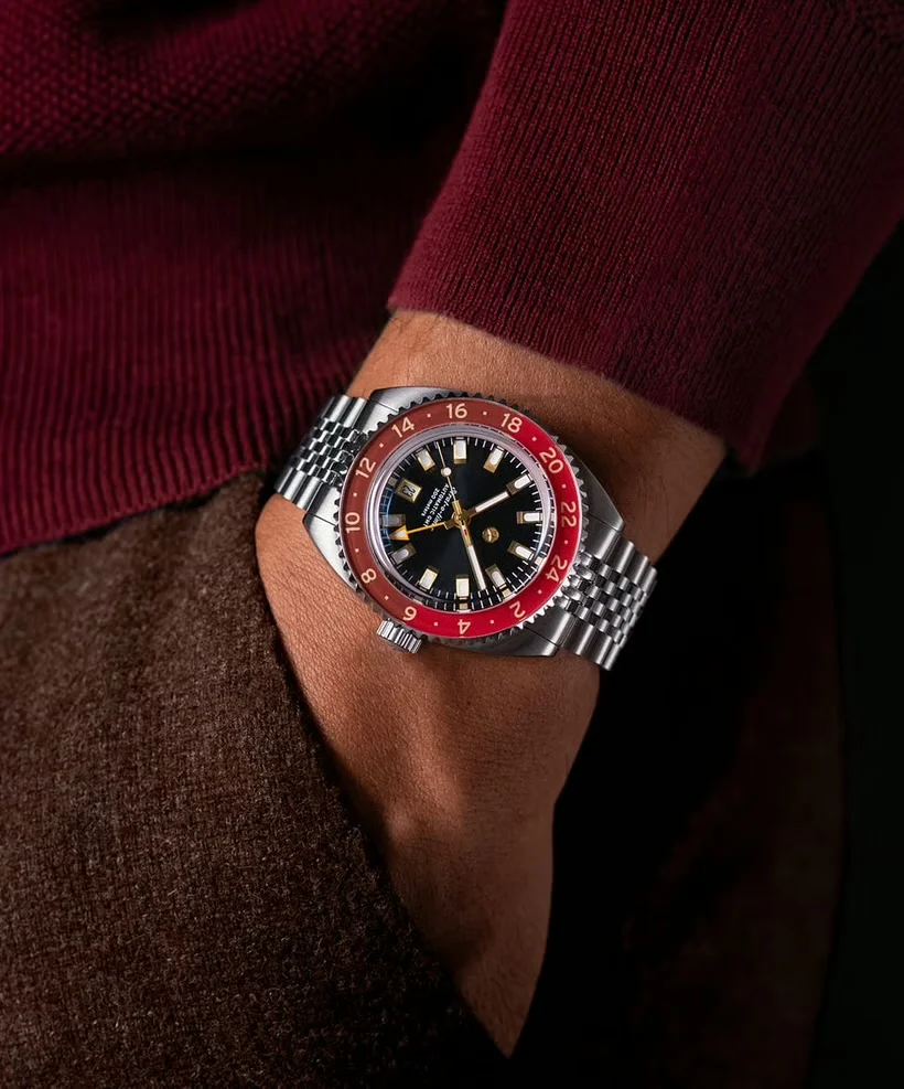 Wrist wearing steel jubilee watch with black dial, gold markers, red/brown GMT bezel. Maroon sweater & brown pants.