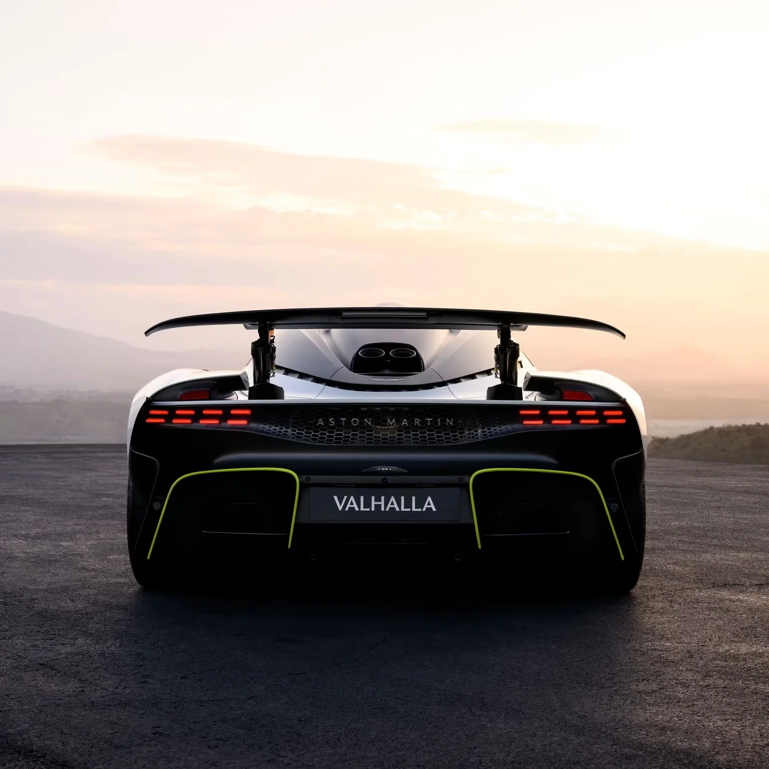 Black Aston Martin Valhalla hypercar's rear with bright green trim, large spoiler, and red LED lights at sunset.