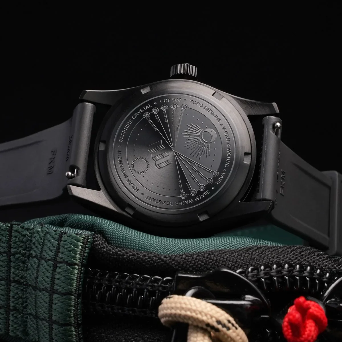 Luxury black watch with intricate engraved case back (sun/moon design, '1 OF 500'), black rubber strap, on a green tactical pouch.