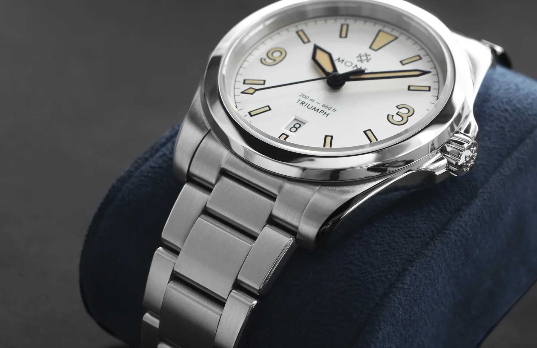 Monta Triumph steel watch with cream dial, yellow 3 & 9 numerals, luminous markers, and bracelet on a blue cushion.