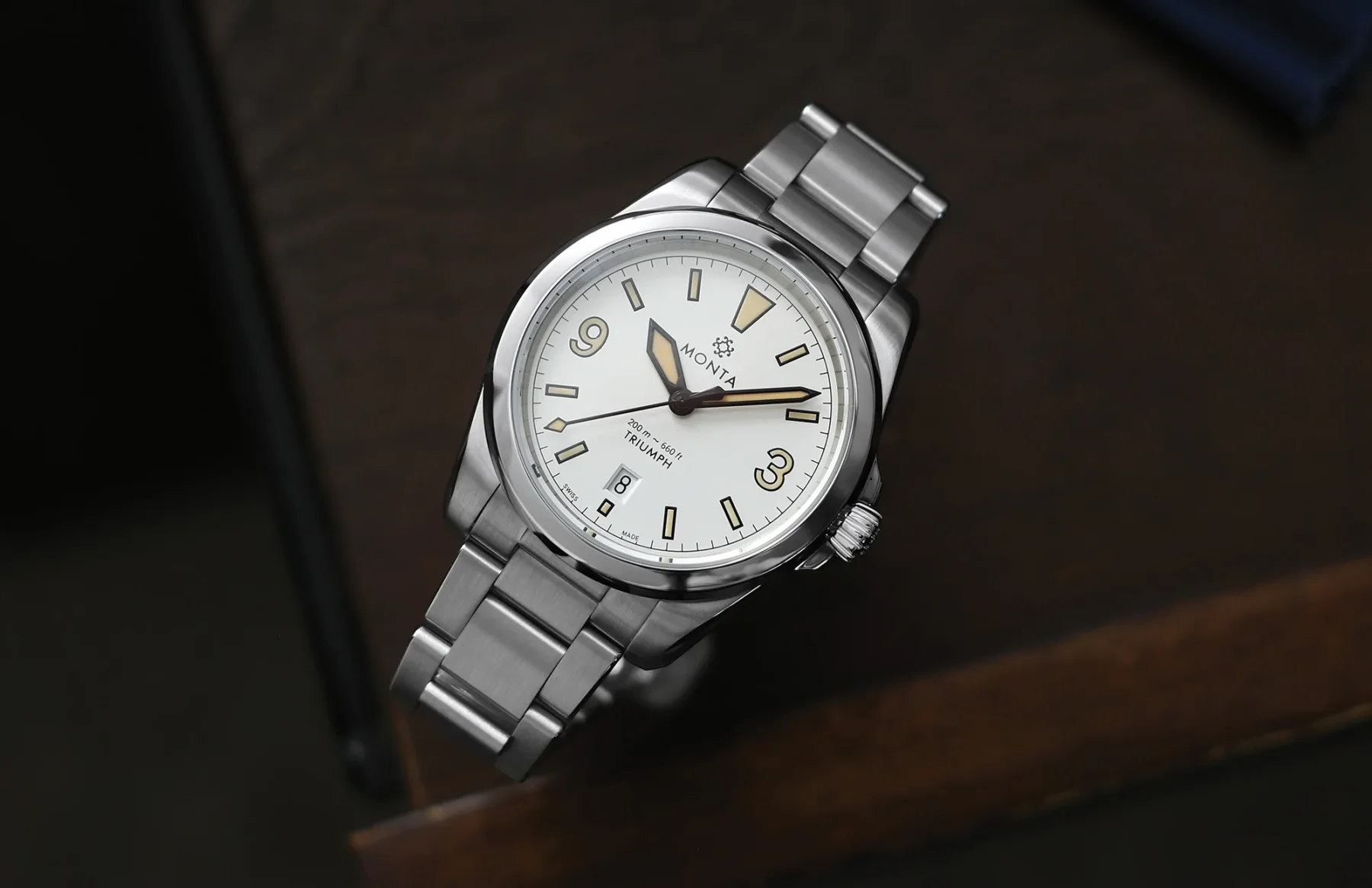 Monta Triumph watch: white dial, steel case & bracelet, yellow-lumed hands/indices, date at 6 o'clock, on wood.