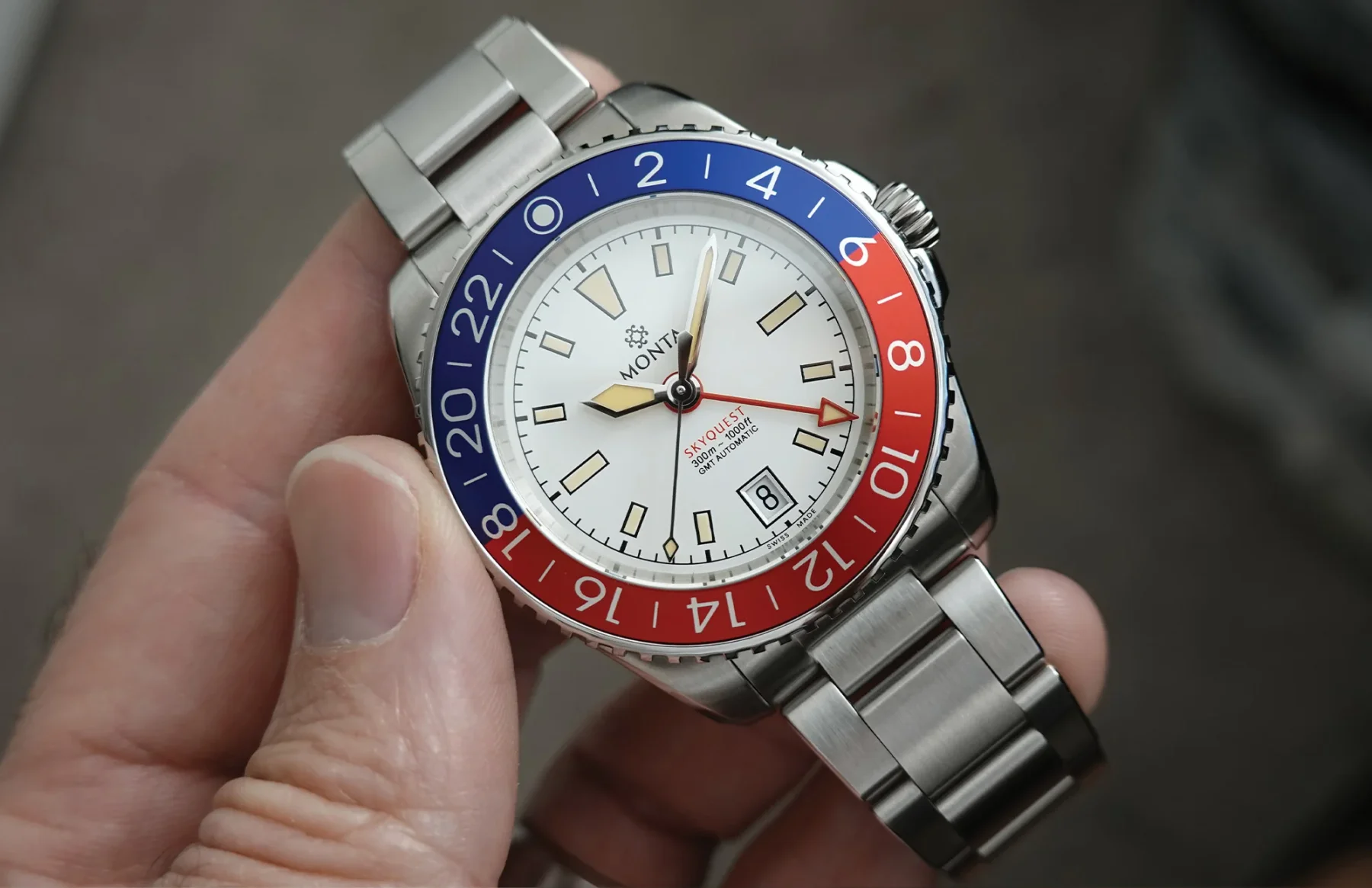Close-up of a Monta Skyquest GMT watch with white dial, steel bracelet, and red and blue 24-hour bezel, held by a hand.