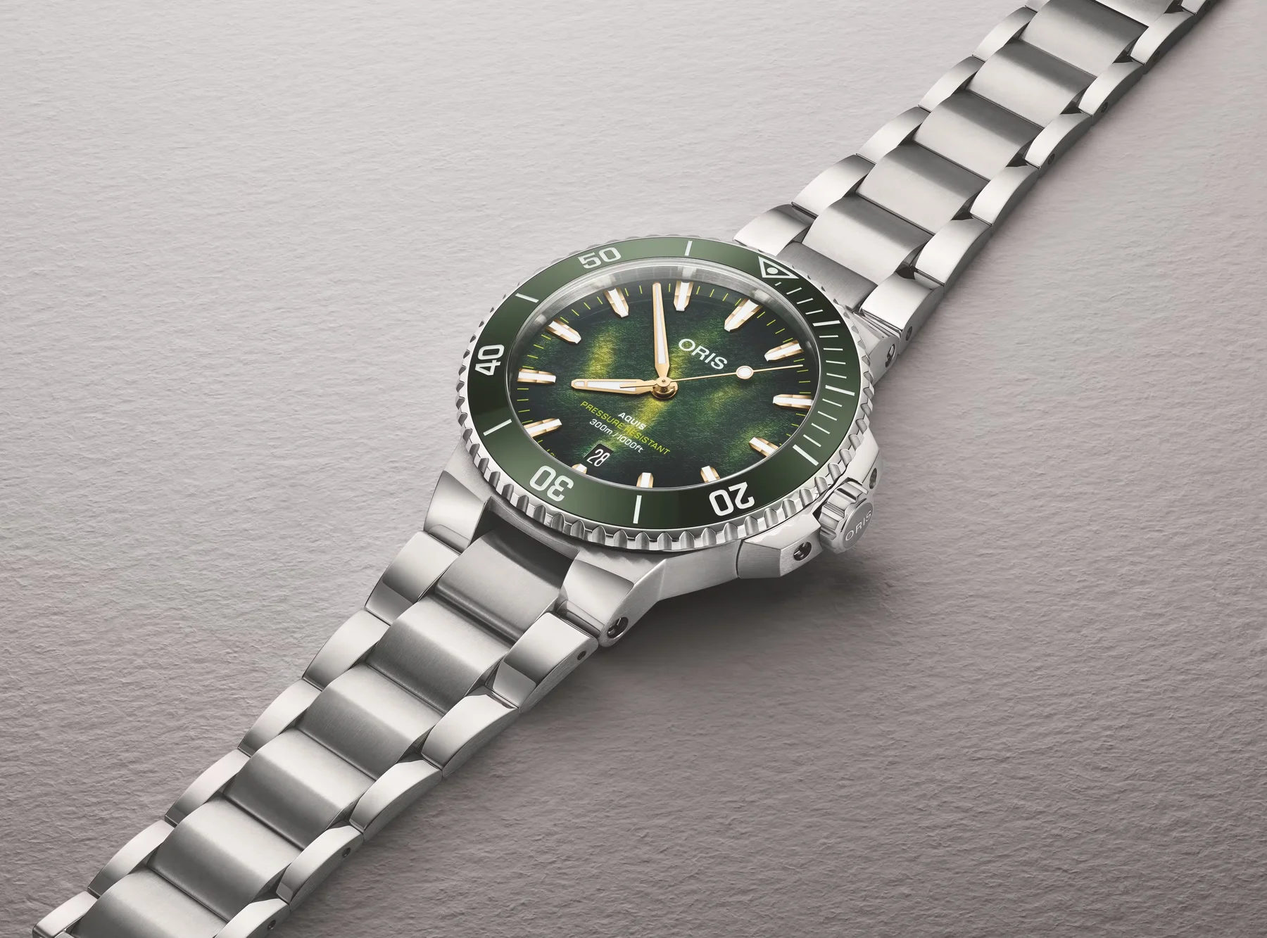 Oris Aquis diver with textured green dial, gold hands, green bezel, date, and steel bracelet on grey.