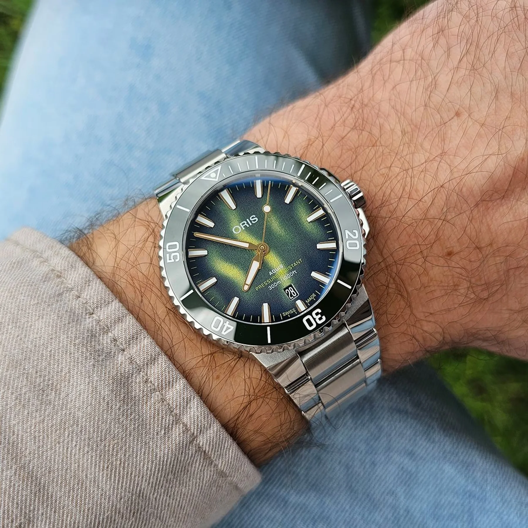 Oris Aquis watch with green-blue gradient dial, date, and steel bracelet on a hairy arm, with denim and blurred green.