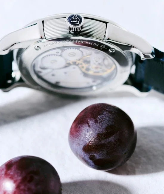 Luxury watch with royal crown on side, exhibition caseback revealing gears, and two purple grapes.