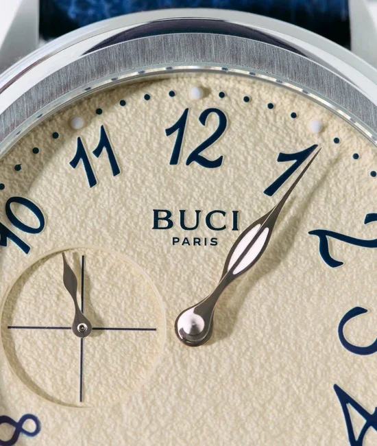 Close-up: Buci Paris watch with textured cream dial, blue Arabic numerals, leaf hands, small seconds sub-dial, brushed bezel.