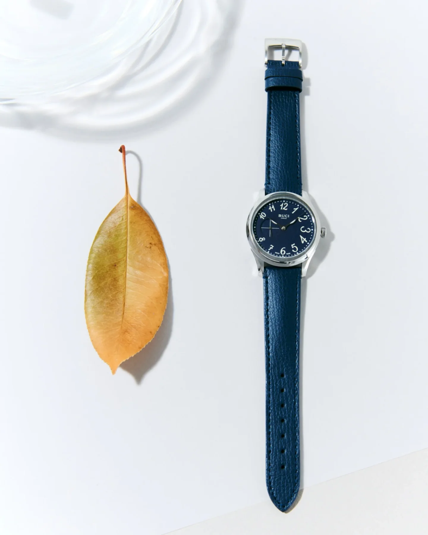 BUCI PARIS watch: blue dial, silver case, textured blue leather strap, next to a faded yellow leaf on white.
