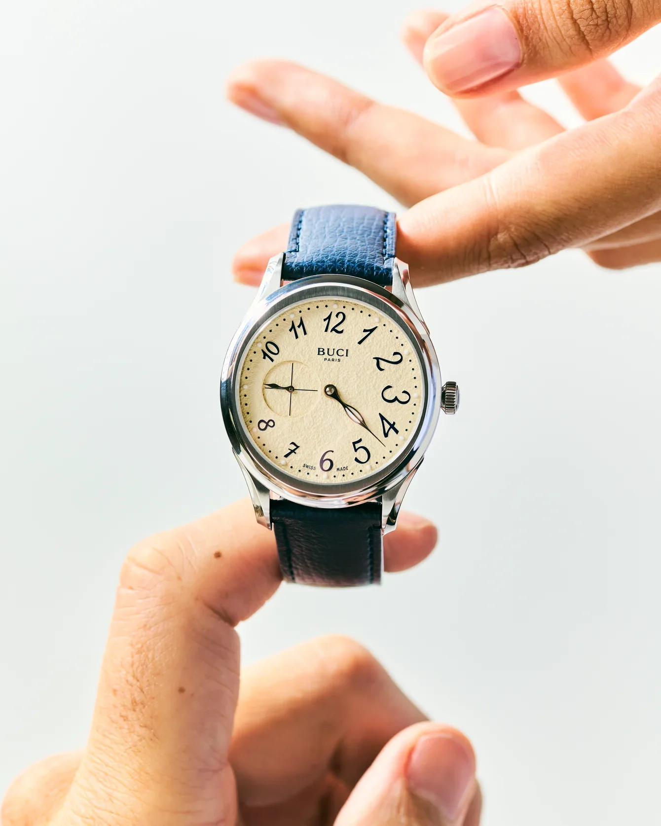 Two hands hold a silver Buci Paris watch with a cream dial, black Arabic numerals, small seconds, and a blue leather strap.