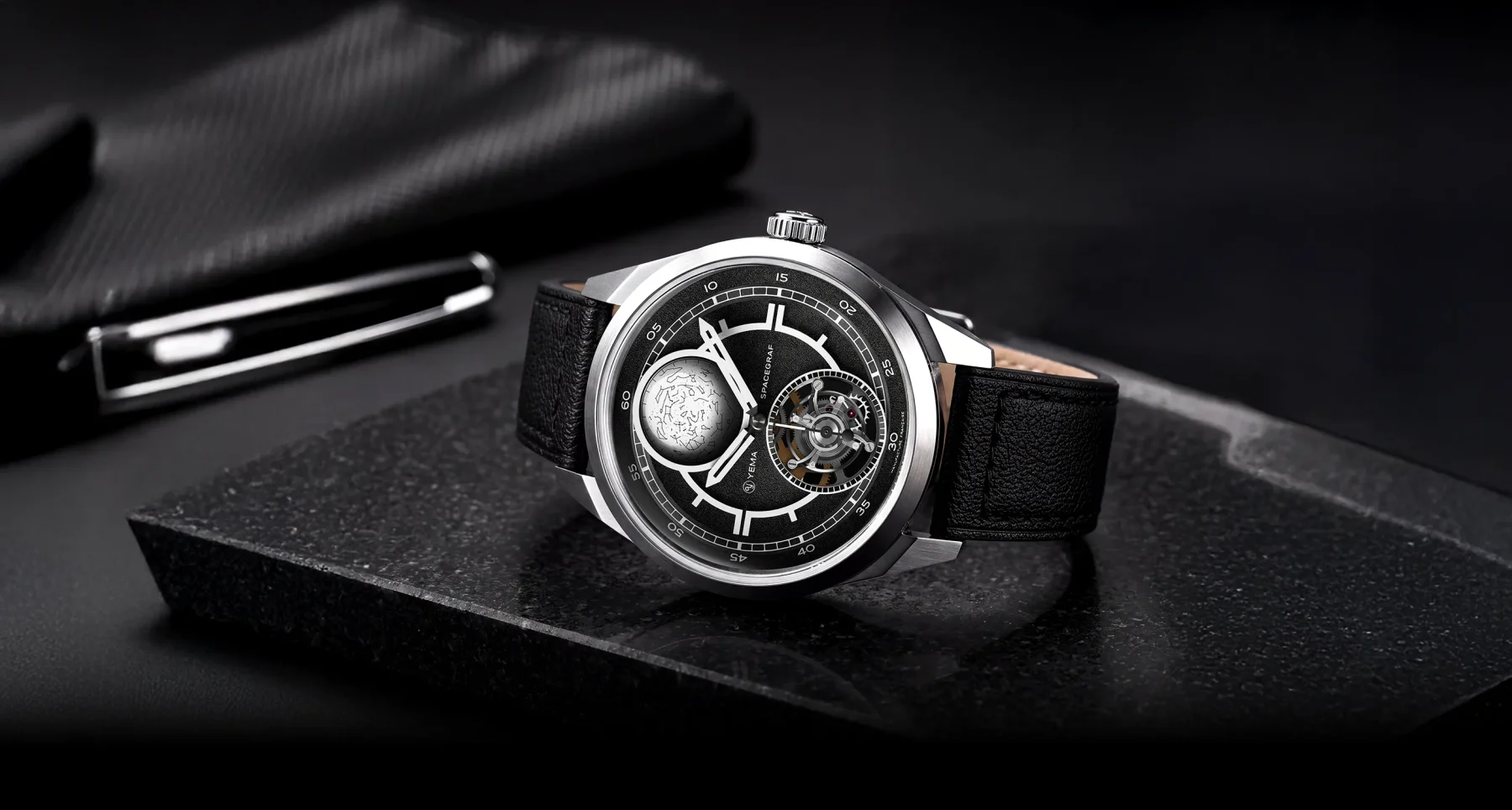 Silver watch with black dial, moonphase, exposed gears, black leather strap, on dark stone. Blurry pen and pouch in background.