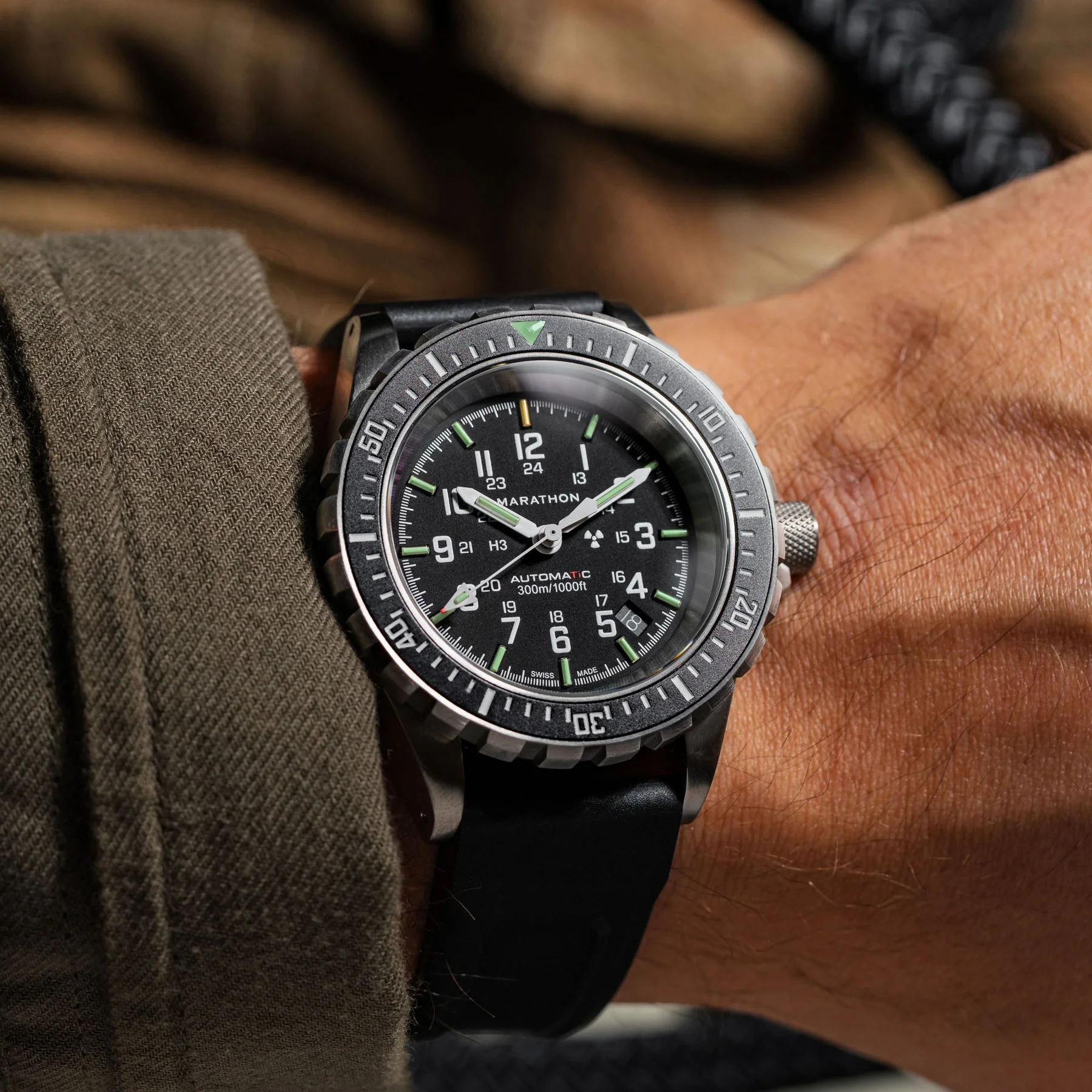 Close-up of a Marathon tactical watch with a black dial, green lume, and grey bezel on a wrist with a brown sleeve.