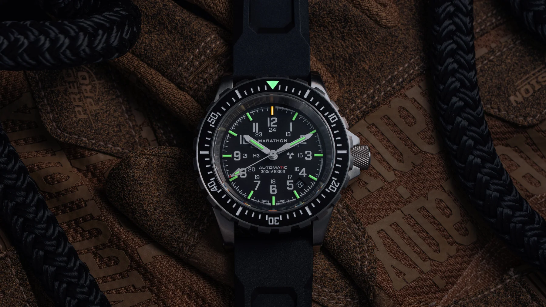 Marathon dive watch with black dial, green luminous markers, black rubber strap, on a brown tactical glove with a braided rope.