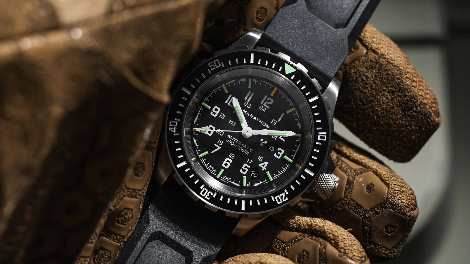 Close-up of a Marathon automatic dive watch with black dial, green lume, steel case, black rubber strap, on a tactical glove.