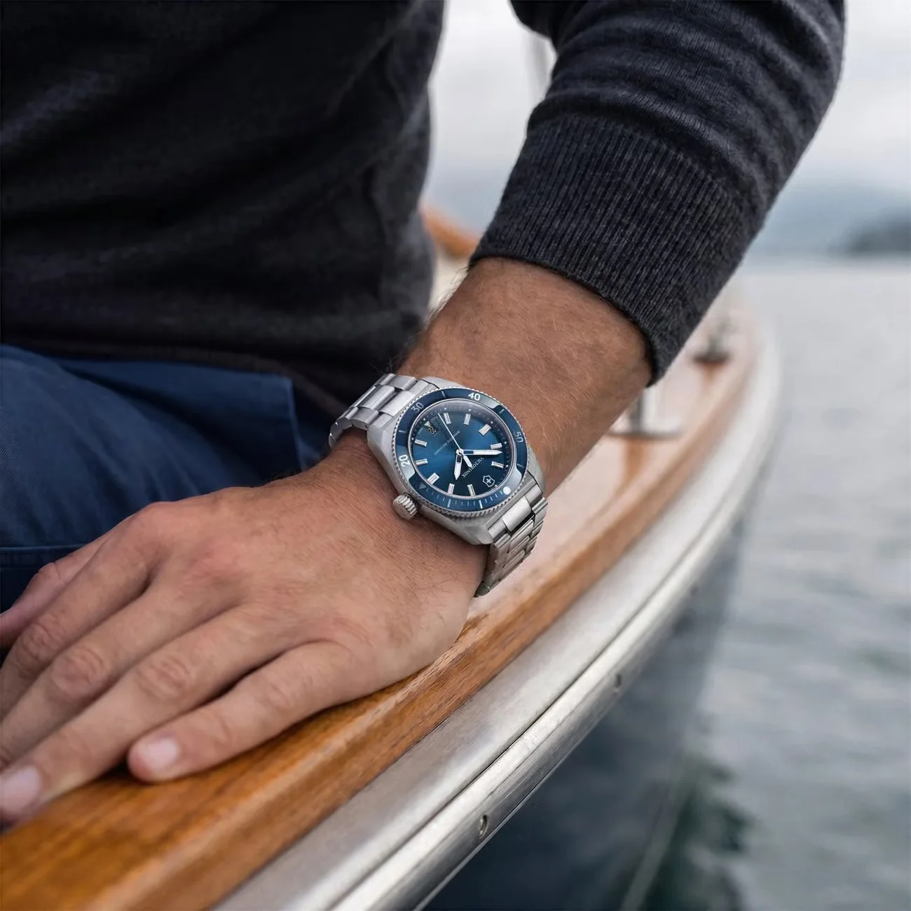 Man's wrist with Victorinox Swiss Army dive watch, blue dial/bezel, steel bracelet, on boat railing overlooking water.