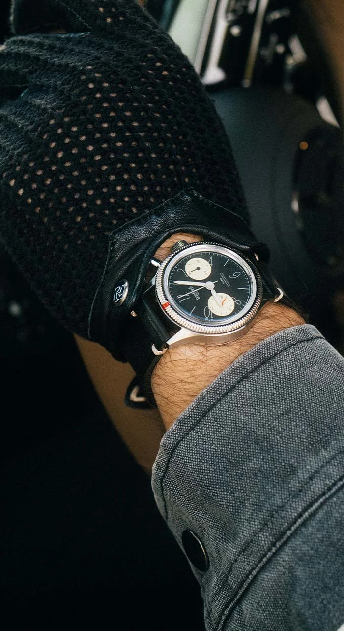 Man's wrist with a Hanhart steel chronograph watch, black dial, cream subdials, fluted bezel. He wears a perforated driving glove & denim jacket.
