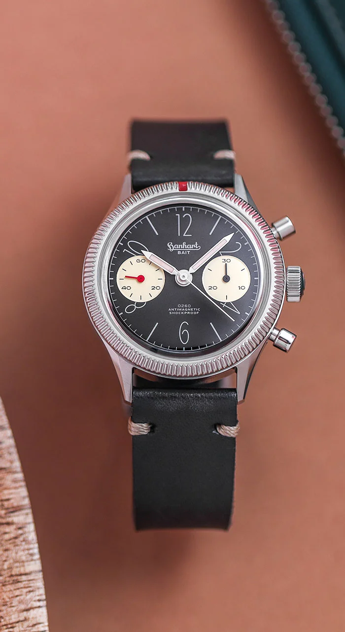 Hanhart 0260 x BAIT chronograph with black dial, cream sub-dials, coin-edge bezel, red marker, & black leather strap on brown surface.