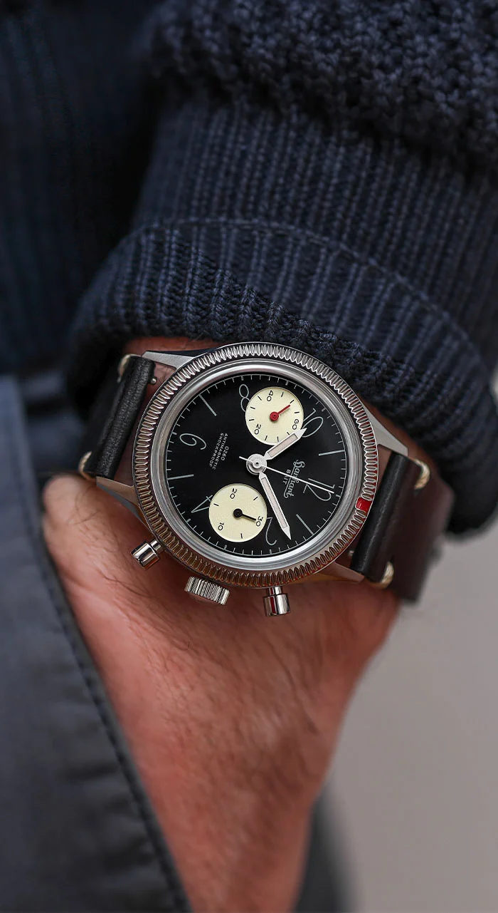 Close-up of a Hanhart 0260 x BAIT chronograph on a wrist, featuring a black dial, cream sub-dials, fluted bezel, and brown leather strap.