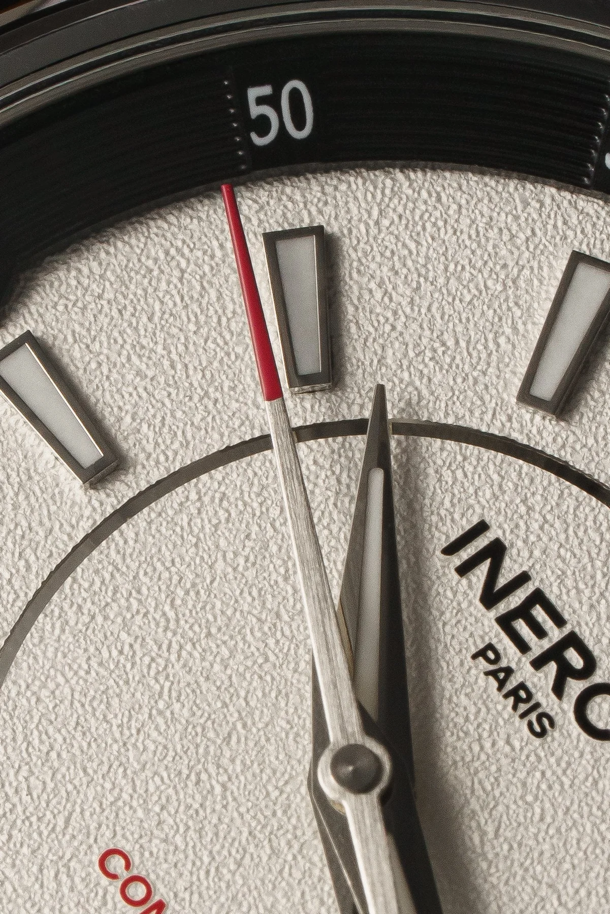 Macro shot of a luxury watch dial: textured white surface, red-tipped seconds hand, sword hand, INEROC PARIS, and 50 on bezel.