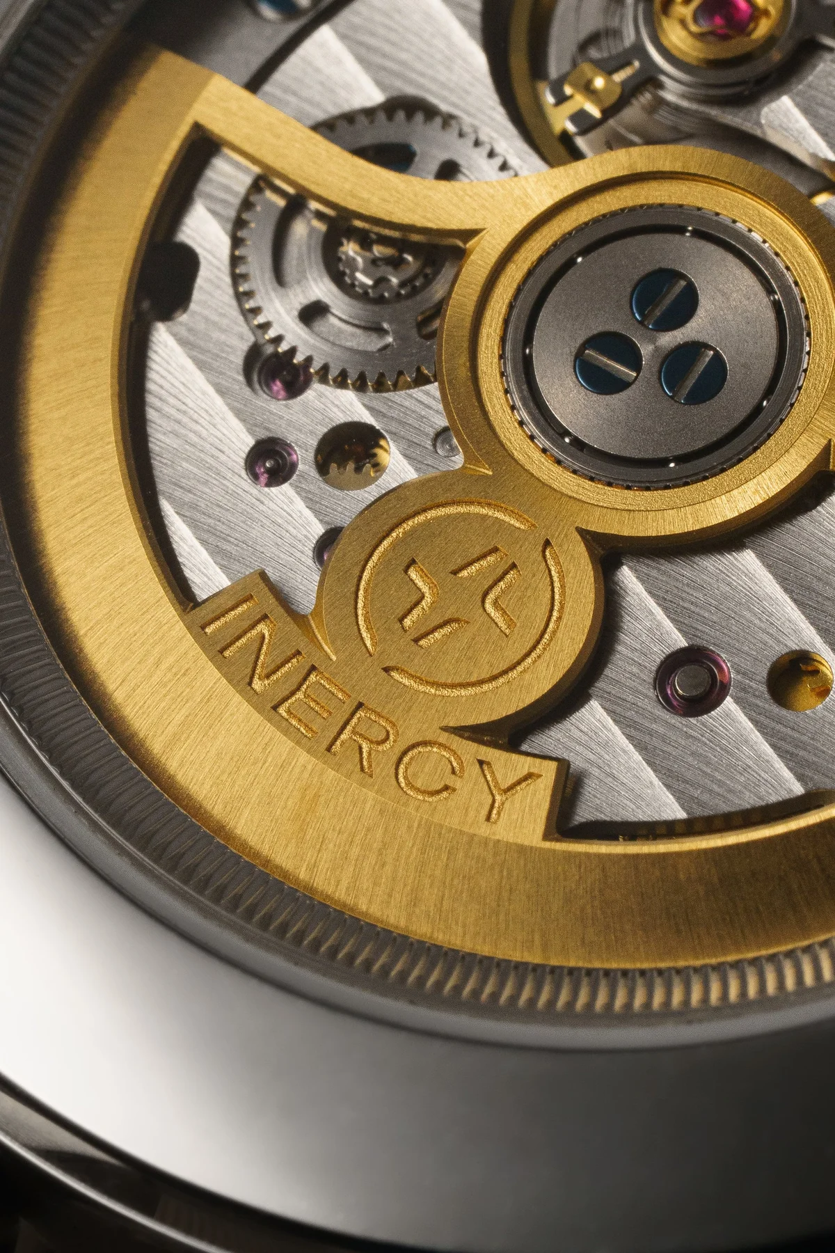 Macro view of a watch movement: gold rotor segment engraved 'INERCY', logo, blued screws, gears, ruby jewels, Geneva stripes.