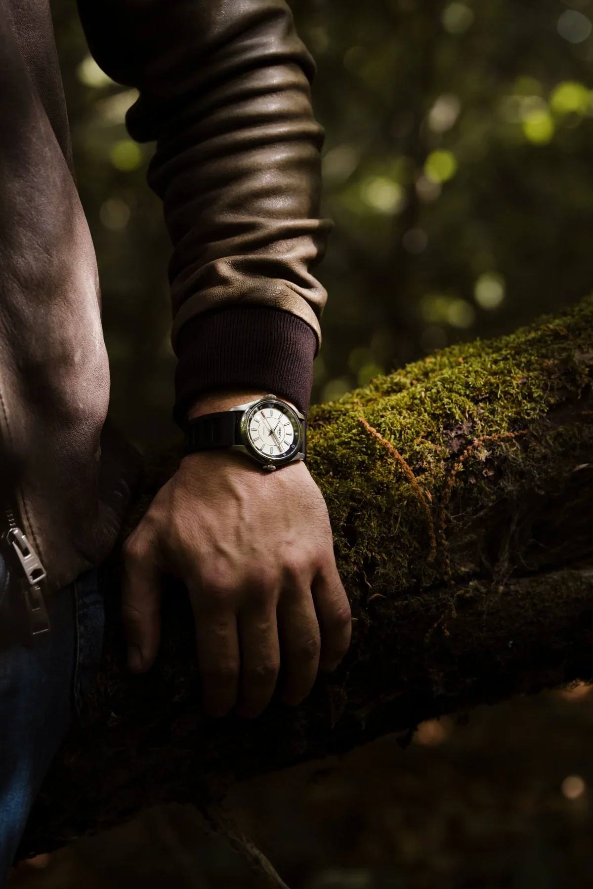 Man in leather jacket wears a cream dial Inercy dual-crown watch, hand resting on a mossy tree branch.