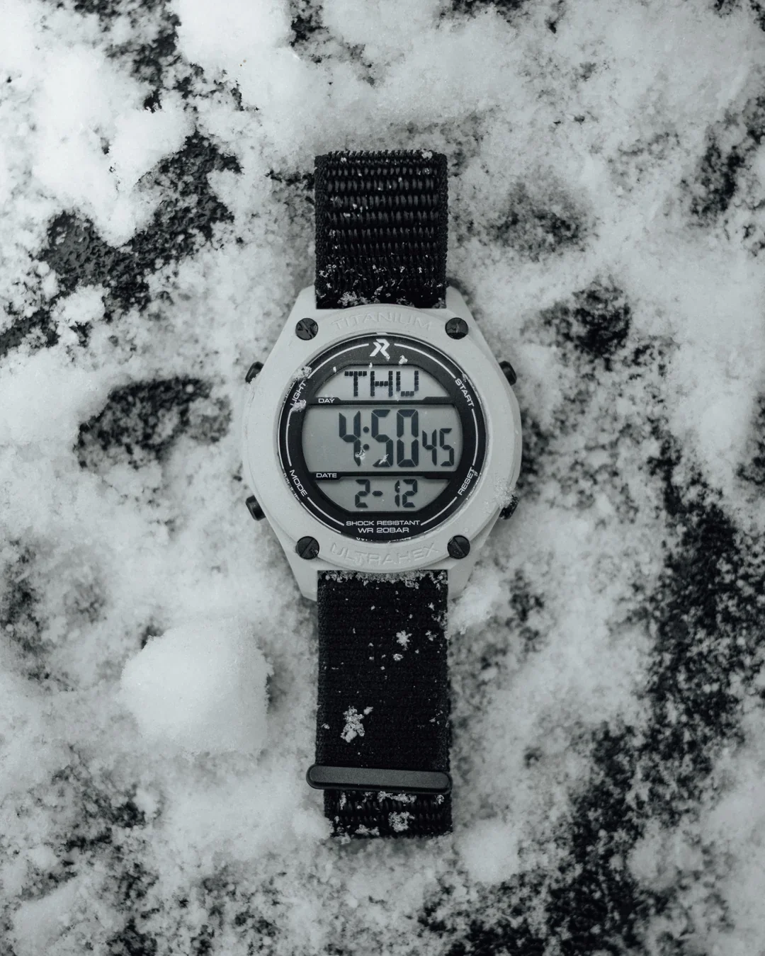 White digital watch with black NATO strap on snow-covered dark ground. Display reads THU 4:50:45, 2-12. Titanium.