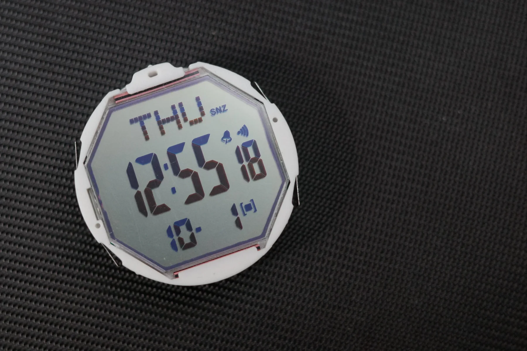 White octagonal digital watch module displaying THU 12:55, 18, 10, and icons, on a dark textured surface.