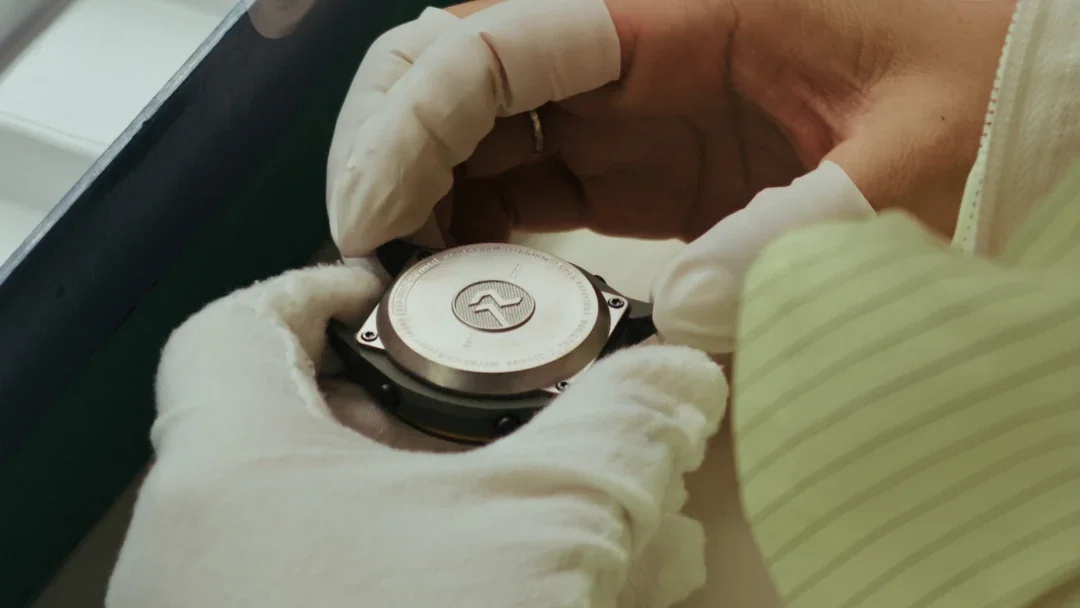 Gloved hands carefully assemble a luxury watch, revealing its engraved metallic case back with a brand logo.
