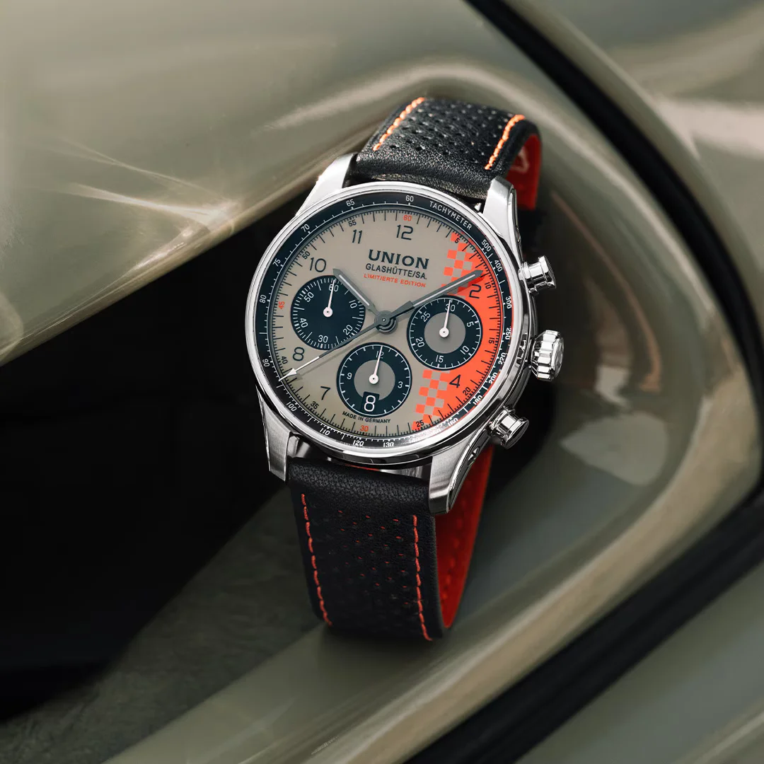 Union Glashütte chronograph watch with beige and orange checkered dial, black perforated strap on a curved, muted green surface.